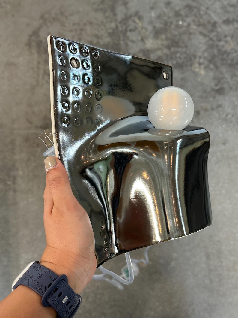K&ouml;nnte beinhalten: Eine Wandleuchte aus Keramik mit gl&auml;nzendem, dunkelmetallischem Finish und einem strukturierten Gittermuster im oberen Bereich. Die Lampe hat eine geschwungene, skulpturale Form und ist mit einer runden, wei&szlig;en Gl&uuml;hbirne aus Glas versehen.