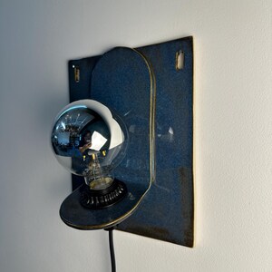 May include: A wall-mounted lamp featuring a rectangular blue ceramic backplate with rounded edges and two small rectangular cutouts at the top. A circular ceramic shelf extends from the center, holding a black socket and a large, reflective silver globe bulb.