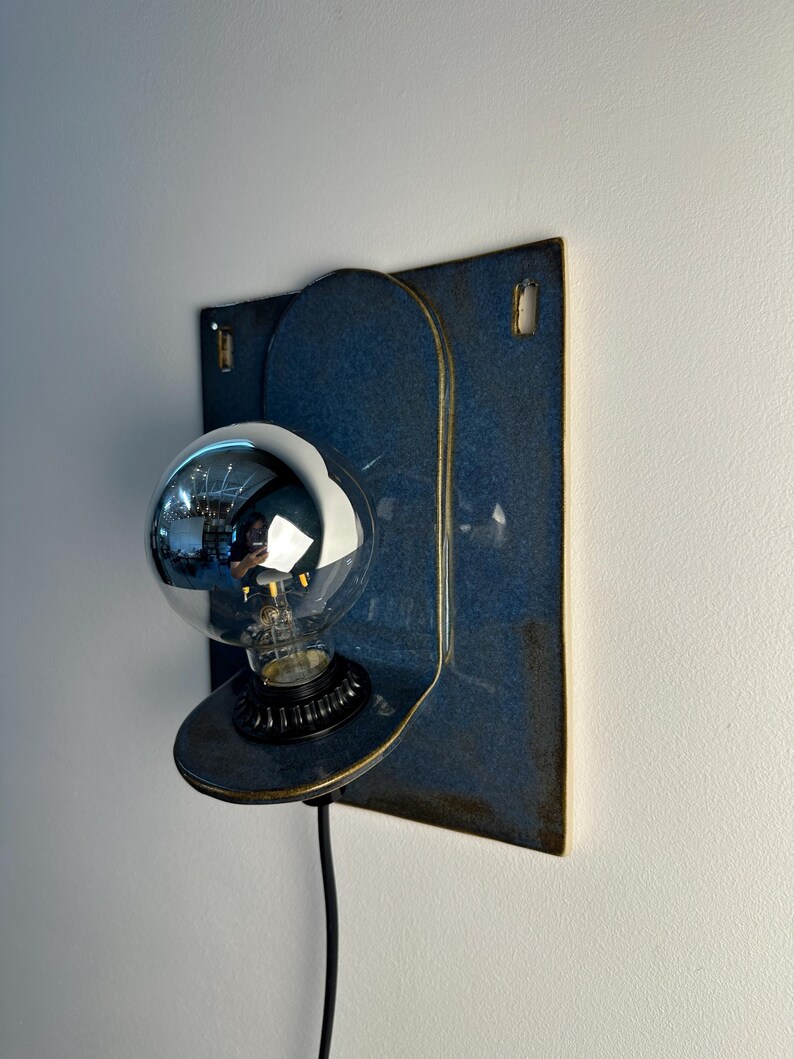 May include: A wall-mounted lamp featuring a rectangular blue ceramic backplate with rounded edges and two small rectangular cutouts at the top. A circular ceramic shelf extends from the center, holding a black socket and a large, reflective silver globe bulb.