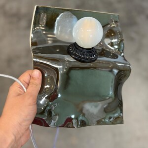 May include: A wall-mounted lamp featuring a glossy, dark green, irregularly shaped ceramic base with a wavy, reflective surface. A round, white, frosted glass light bulb is attached to the center of the ceramic piece via a black socket.