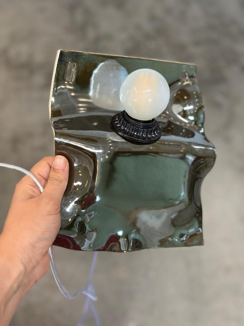 May include: A wall-mounted lamp featuring a glossy, dark green, irregularly shaped ceramic base with a wavy, reflective surface. A round, white, frosted glass light bulb is attached to the center of the ceramic piece via a black socket.