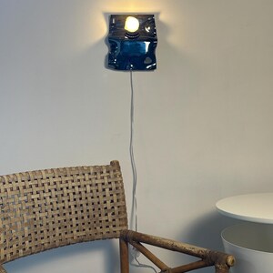 May include: A wall-mounted lamp featuring a glossy, dark blue, wavy ceramic base with a single exposed light bulb emitting a warm glow. A thin white power cord hangs vertically from the base of the fixture down the wall toward the floor.