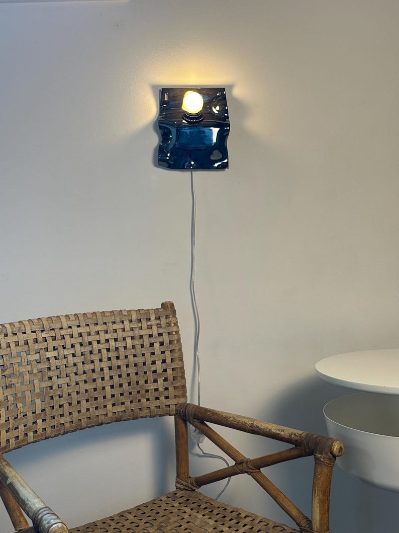 May include: A wall-mounted lamp featuring a glossy, dark blue, wavy ceramic base with a single exposed light bulb emitting a warm glow. A thin white power cord hangs vertically from the base of the fixture down the wall toward the floor.