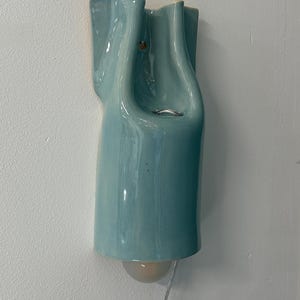 May include: A wall-mounted light fixture made of glossy, light blue ceramic with a fluid, organic shape. The fixture features a curved, vertical design with a small circular opening near the top and a single light bulb extending from the bottom.