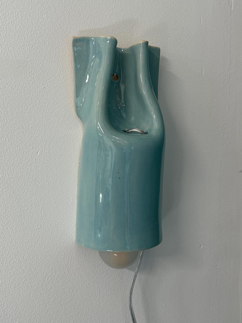 May include: A wall-mounted light fixture made of glossy, light blue ceramic with a fluid, organic shape. The fixture features a curved, vertical design with a small circular opening near the top and a single light bulb extending from the bottom.