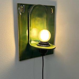 May include: A wall-mounted ceramic sconce featuring a glossy, deep green rectangular backplate with an arched center section. A small, round, frosted glass light bulb sits on a protruding shelf, connected to a black cord hanging downward.