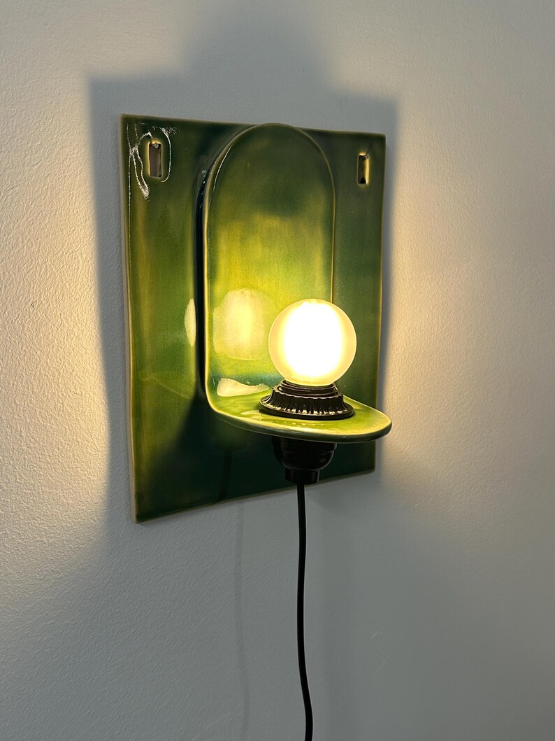 May include: A wall-mounted ceramic sconce featuring a glossy, deep green rectangular backplate with an arched center section. A small, round, frosted glass light bulb sits on a protruding shelf, connected to a black cord hanging downward.