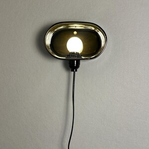 K&ouml;nnte beinhalten: Eine ovale Wandleuchte mit schwarzem Rahmen und goldfarbenem Innenraum. Die Leuchte beherbergt eine helle, runde Gl&uuml;hbirne. Ein schwarzes Kabel erstreckt sich nach unten von der Leuchte.