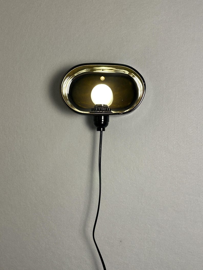 K&ouml;nnte beinhalten: Eine ovale Wandleuchte mit schwarzem Rahmen und goldfarbenem Innenraum. Die Leuchte beherbergt eine helle, runde Gl&uuml;hbirne. Ein schwarzes Kabel erstreckt sich nach unten von der Leuchte.