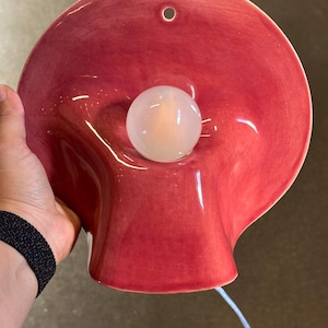 May include: A wall-mounted ceramic lamp with a glossy, deep red, bowl-shaped shade. The lamp features a central, spherical white light bulb and a small circular hole at the top for mounting. A thin white electrical cord extends from the bottom.