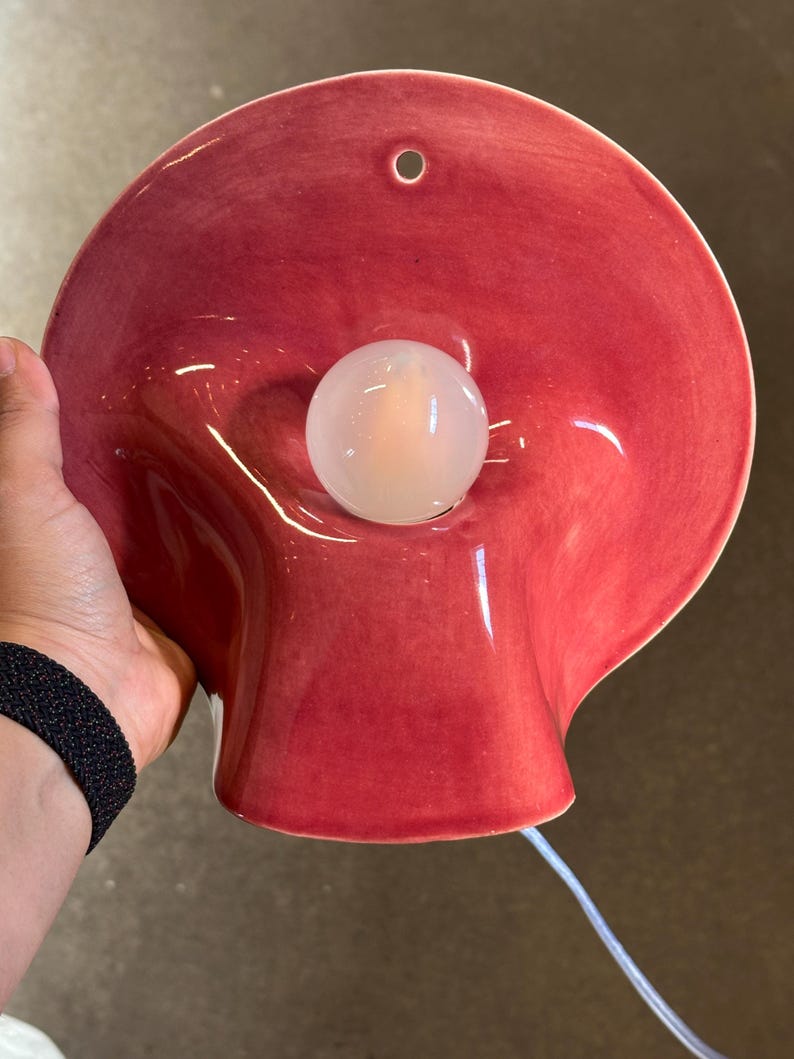 May include: A wall-mounted ceramic lamp with a glossy, deep red, bowl-shaped shade. The lamp features a central, spherical white light bulb and a small circular hole at the top for mounting. A thin white electrical cord extends from the bottom.