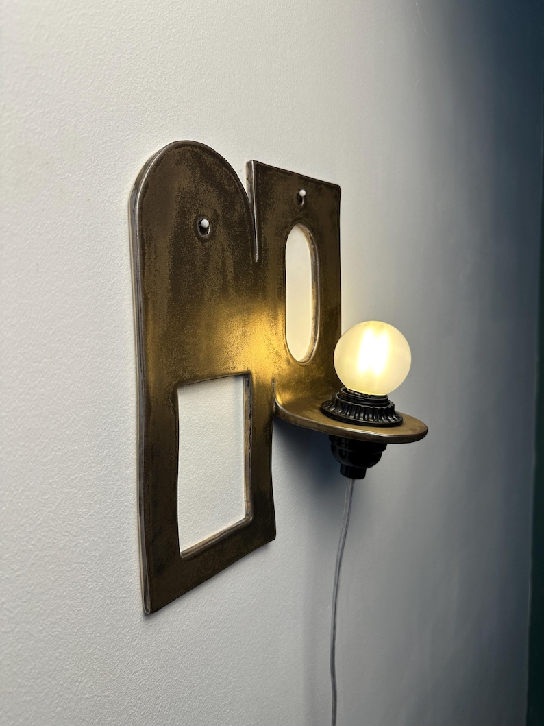 K&ouml;nnte beinhalten: Eine Wandleuchte mit einer dunklen, metallischen, bogenf&ouml;rmigen R&uuml;ckplatte, die einen rechteckigen Ausschnitt und eine ovale &Ouml;ffnung aufweist. Ein kleines, kreisf&ouml;rmiges Regal ragt an der rechten Seite hervor und tr&auml;gt eine leuchtende, mattierte Kugel.