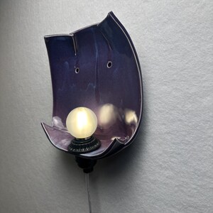 May include: A wall-mounted lamp featuring a curved, glossy purple ceramic shade with two small circular holes near the top. A round, frosted white light bulb is set into a black base at the bottom of the ceramic piece, with a thin power cord extending downwards.