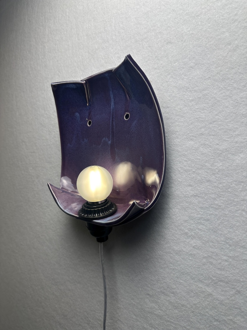 May include: A wall-mounted lamp featuring a curved, glossy purple ceramic shade with two small circular holes near the top. A round, frosted white light bulb is set into a black base at the bottom of the ceramic piece, with a thin power cord extending downwards.