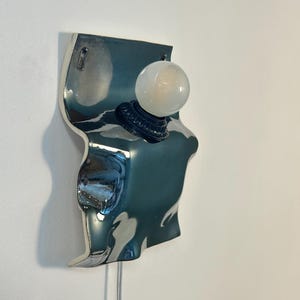 May include: A wall-mounted lamp featuring a reflective, metallic silver, wavy-shaped ceramic base. A dark circular socket is mounted to the center of the base, holding a single, round, frosted white glass light bulb. A thin white cord hangs below.