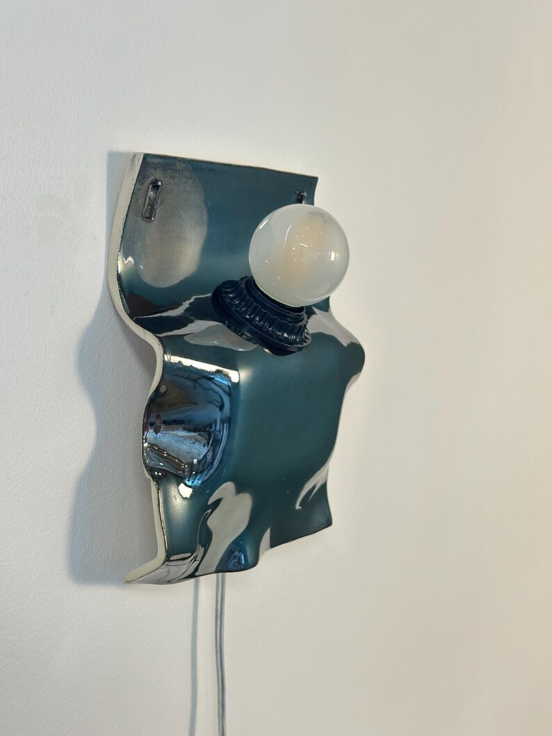 May include: A wall-mounted lamp featuring a reflective, metallic silver, wavy-shaped ceramic base. A dark circular socket is mounted to the center of the base, holding a single, round, frosted white glass light bulb. A thin white cord hangs below.