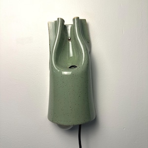May include: A wall-mounted ceramic light fixture in a sage green color with a speckled finish. The lamp features a vertical, elongated shape with a central opening and a black power cord extending from the bottom, showcasing a smooth, glossy glaze.