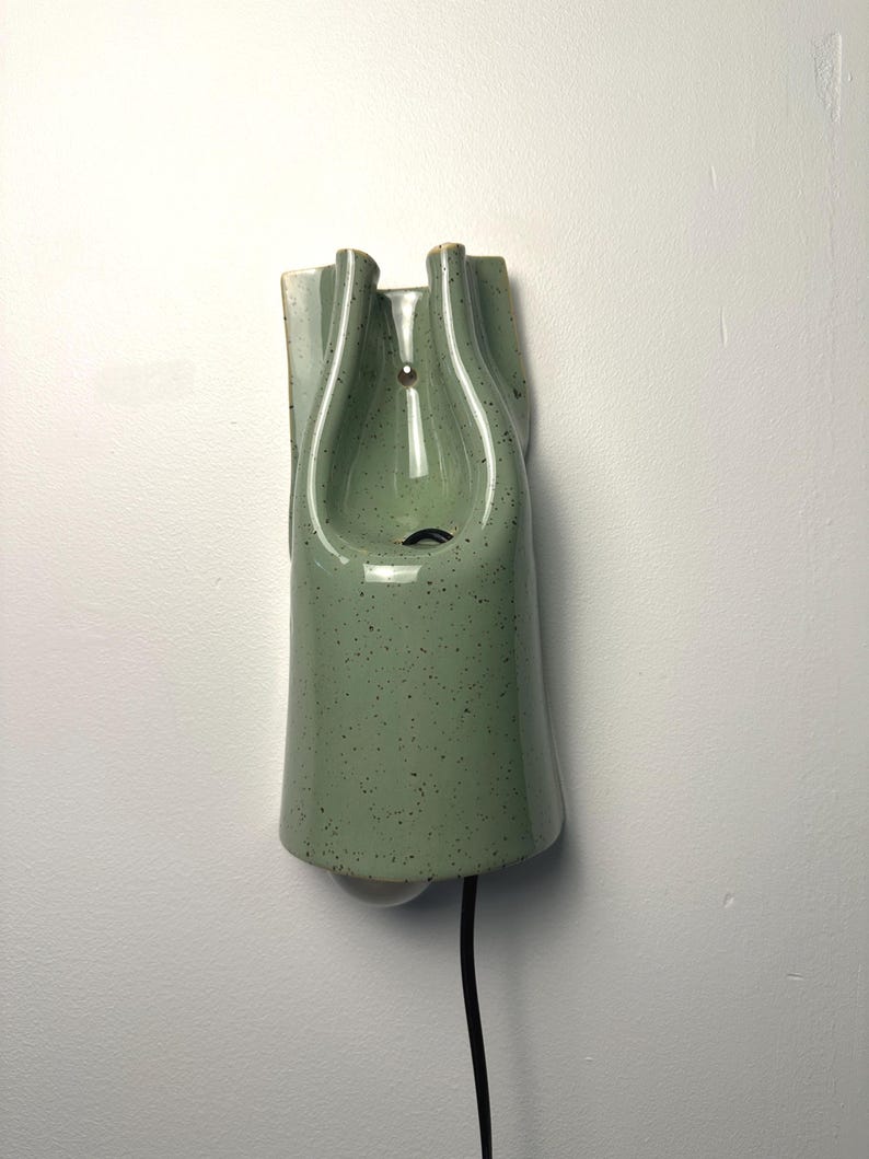 May include: A wall-mounted ceramic light fixture in a sage green color with a speckled finish. The lamp features a vertical, elongated shape with a central opening and a black power cord extending from the bottom, showcasing a smooth, glossy glaze.