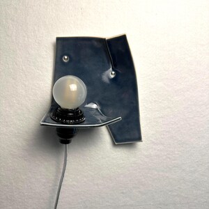 May include: A modern wall sconce with a dark blue ceramic shade and a round, frosted light bulb. The sconce has a black base and a silver cord. The ceramic shade has a unique, asymmetrical shape and is mounted on a white wall.
