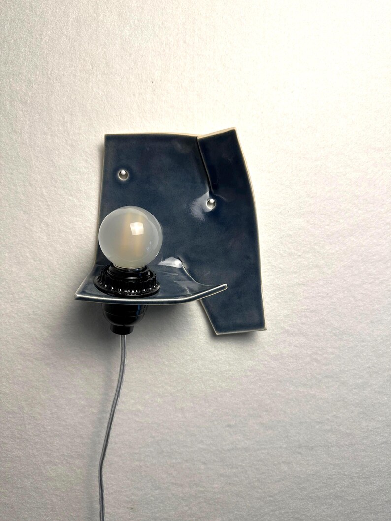 May include: A modern wall sconce with a dark blue ceramic shade and a round, frosted light bulb. The sconce has a black base and a silver cord. The ceramic shade has a unique, asymmetrical shape and is mounted on a white wall.
