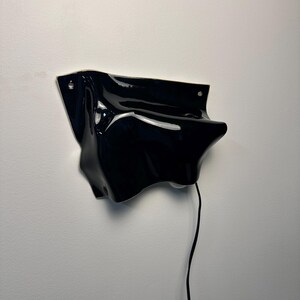 Puede incluir: Una l&aacute;mpara de pared con una forma fluida de cer&aacute;mica o pl&aacute;stico negro brillante de contornos irregulares. La estructura tiene dos orificios de montaje visibles en las esquinas superiores y un cable el&eacute;ctrico negro fino que cuelga con un interruptor.