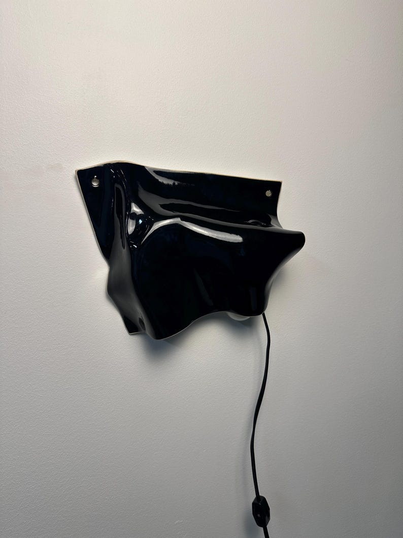 Puede incluir: Una l&aacute;mpara de pared con una forma fluida de cer&aacute;mica o pl&aacute;stico negro brillante de contornos irregulares. La estructura tiene dos orificios de montaje visibles en las esquinas superiores y un cable el&eacute;ctrico negro fino que cuelga con un interruptor.