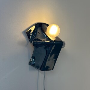 May include: A wall-mounted lamp featuring a glossy, dark navy blue ceramic base with an irregular, folded shape. A single, spherical, warm-toned light bulb is attached to the upper right side of the ceramic form, casting a soft glow against the wall.