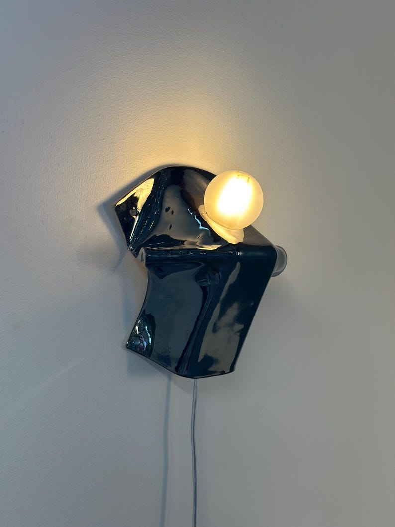May include: A wall-mounted lamp featuring a glossy, dark navy blue ceramic base with an irregular, folded shape. A single, spherical, warm-toned light bulb is attached to the upper right side of the ceramic form, casting a soft glow against the wall.
