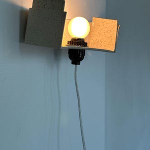 May include: A modern wall sconce with a speckled, light gray geometric design. The sconce features a round, illuminated light bulb and a black socket. A gray electrical cord hangs down from the fixture.