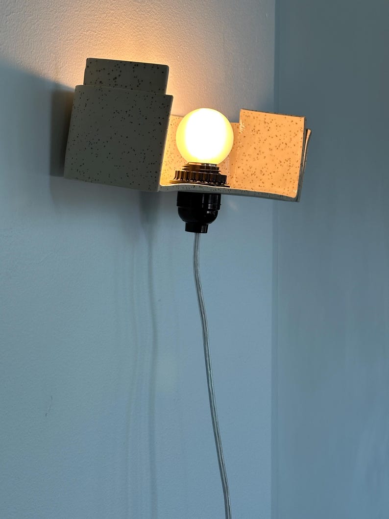 May include: A modern wall sconce with a speckled, light gray geometric design. The sconce features a round, illuminated light bulb and a black socket. A gray electrical cord hangs down from the fixture.