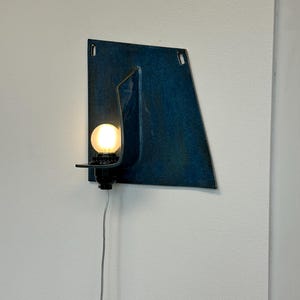 May include: A wall-mounted light fixture with a dark blue, geometric metal shade. The fixture has a small, round light bulb and a black base. A gray power cord extends downward.