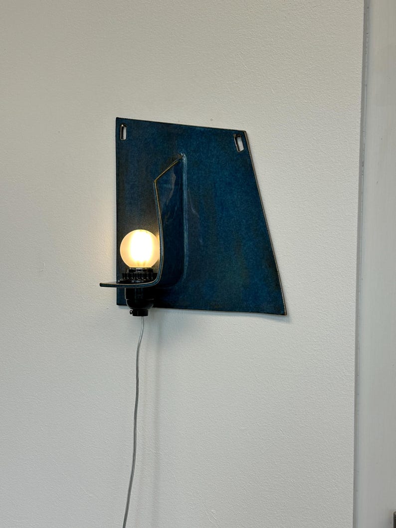 May include: A wall-mounted light fixture with a dark blue, geometric metal shade. The fixture has a small, round light bulb and a black base. A gray power cord extends downward.