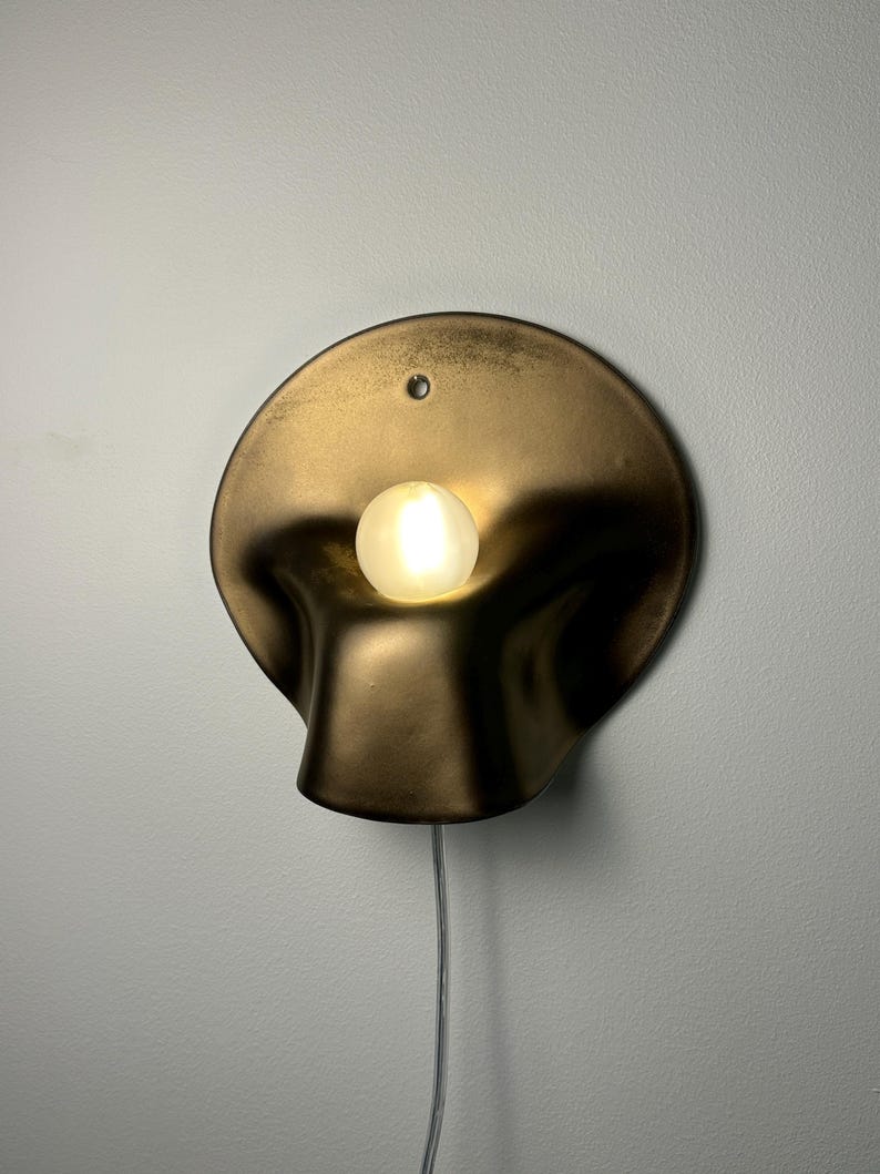May include: A wall-mounted lamp featuring a circular, metallic bronze-toned shade with a fluid, organic, and concave shape. A single, small, spherical frosted light bulb is positioned in the center, emitting a warm glow. A thin, clear power cord hangs below.