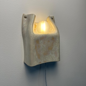 May include: A wall-mounted light fitting with a textured, off-white ceramic housing featuring a U-shaped top opening. A single, glowing, spherical frosted glass bulb sits within the recessed centre, and a thin power cord extends from the bottom.