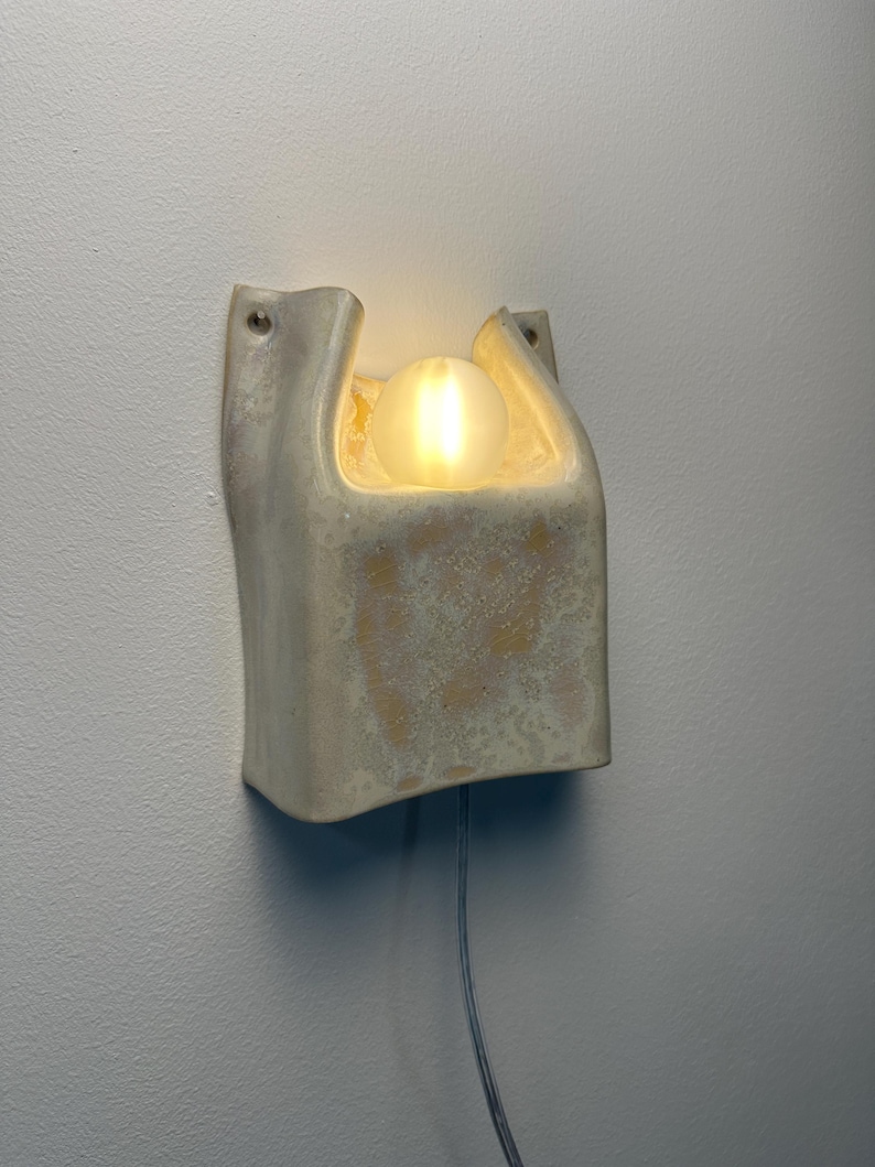 May include: A wall-mounted light fitting with a textured, off-white ceramic housing featuring a U-shaped top opening. A single, glowing, spherical frosted glass bulb sits within the recessed centre, and a thin power cord extends from the bottom.