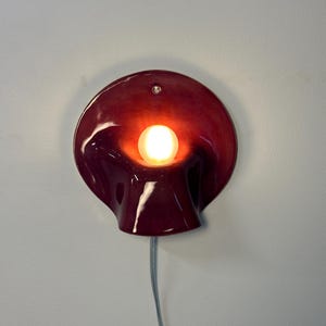 May include: A deep red, glossy ceramic wall sconce with a curved, organic shape resembling a calla lily. The fixture features a central, glowing round light bulb and a small circular mounting hole at the top, with a thin power cord extending downwards.