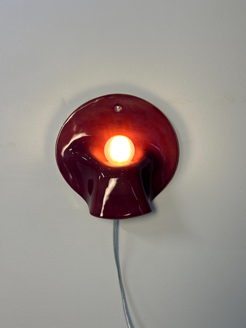 May include: A deep red, glossy ceramic wall sconce with a curved, organic shape resembling a calla lily. The fixture features a central, glowing round light bulb and a small circular mounting hole at the top, with a thin power cord extending downwards.