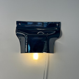 May include: A glossy, dark navy blue ceramic wall sconce with a wide, flared top section and a curved lower housing. The fixture features two small circular mounting holes at the top corners and emits a warm, soft glow from a single light bulb.