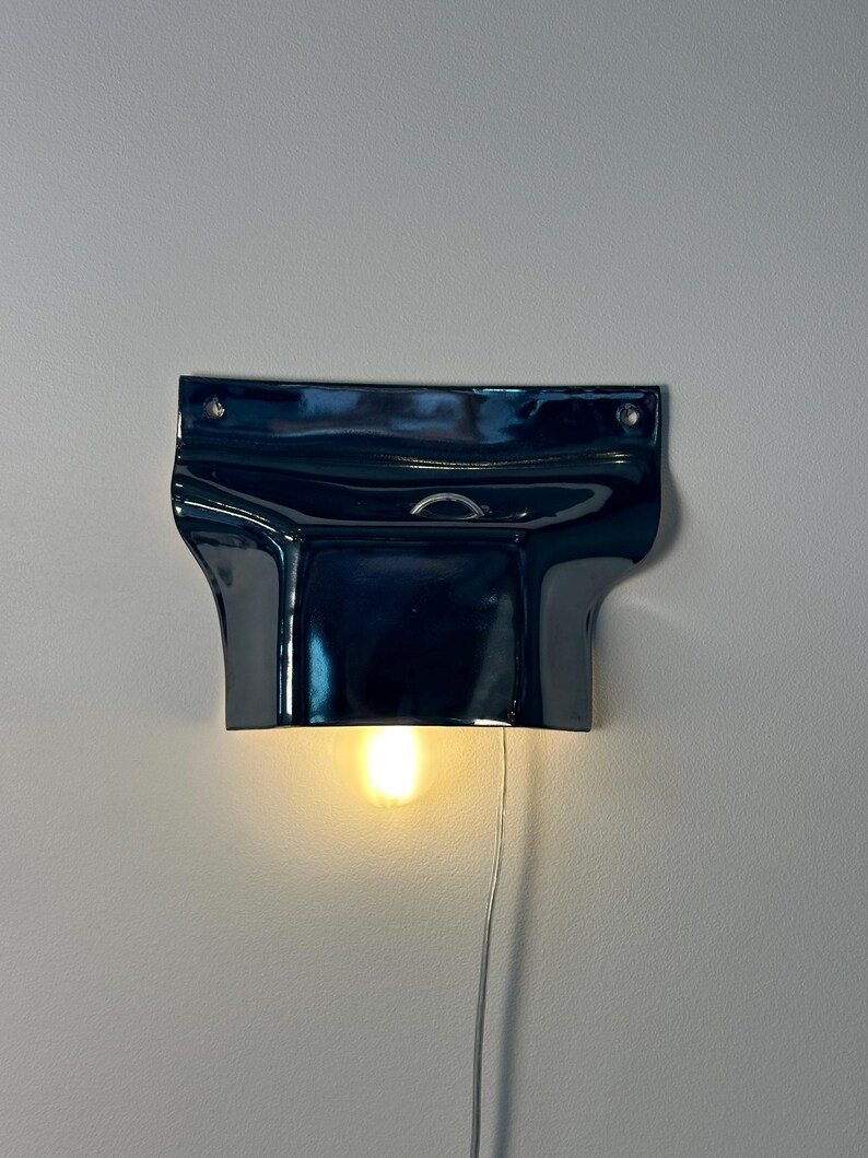 May include: A glossy, dark navy blue ceramic wall sconce with a wide, flared top section and a curved lower housing. The fixture features two small circular mounting holes at the top corners and emits a warm, soft glow from a single light bulb.