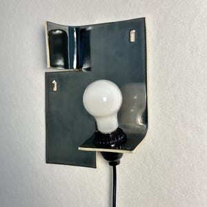 Pode incluir: Uma lumin&aacute;ria de parede moderna com uma l&acirc;mpada branca colocada contra uma placa de metal curva cinza escuro. A placa tem uma borda dourada e &eacute; montada em uma parede branca. A l&acirc;mpada est&aacute; conectada a um cabo preto.
