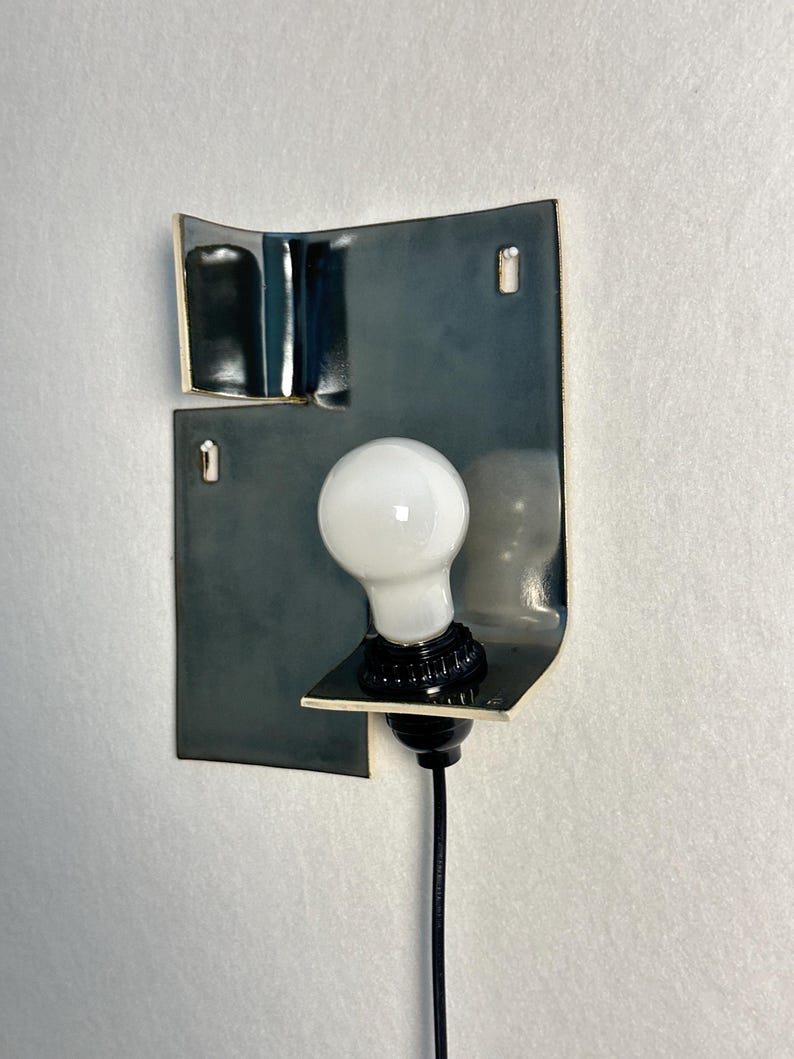 Pode incluir: Uma lumin&aacute;ria de parede moderna com uma l&acirc;mpada branca colocada contra uma placa de metal curva cinza escuro. A placa tem uma borda dourada e &eacute; montada em uma parede branca. A l&acirc;mpada est&aacute; conectada a um cabo preto.
