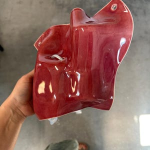 Puede incluir: Una presa de escalada de pared de cer&aacute;mica en color rojo intenso brillante con una forma org&aacute;nica irregular. La pieza presenta una superficie lisa y reflectante con un orificio de montaje circular en la parte superior y un &aacute;rea rebajada.
