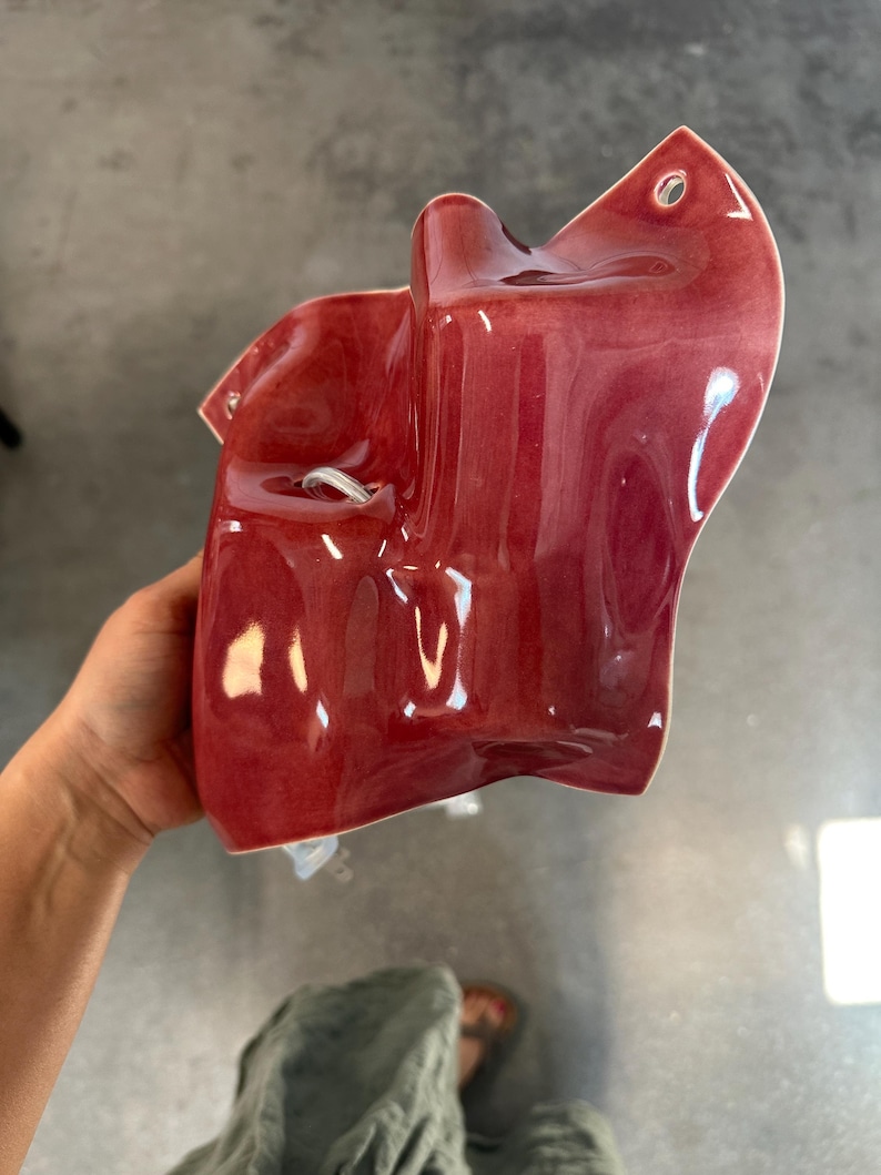 Puede incluir: Una presa de escalada de pared de cer&aacute;mica en color rojo intenso brillante con una forma org&aacute;nica irregular. La pieza presenta una superficie lisa y reflectante con un orificio de montaje circular en la parte superior y un &aacute;rea rebajada.