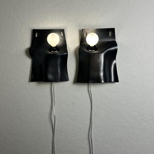 May include: Two black wall sconces with a wavy, sheet-like design, each featuring a single round, glowing white light bulb. The fixtures have rectangular cutouts at the top and thin, clear power cords extending downward from the base of each unit.
