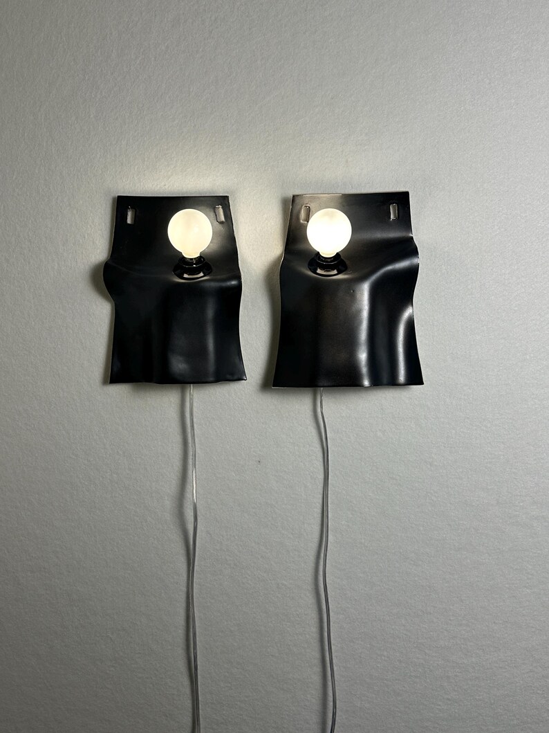 May include: Two black wall sconces with a wavy, sheet-like design, each featuring a single round, glowing white light bulb. The fixtures have rectangular cutouts at the top and thin, clear power cords extending downward from the base of each unit.