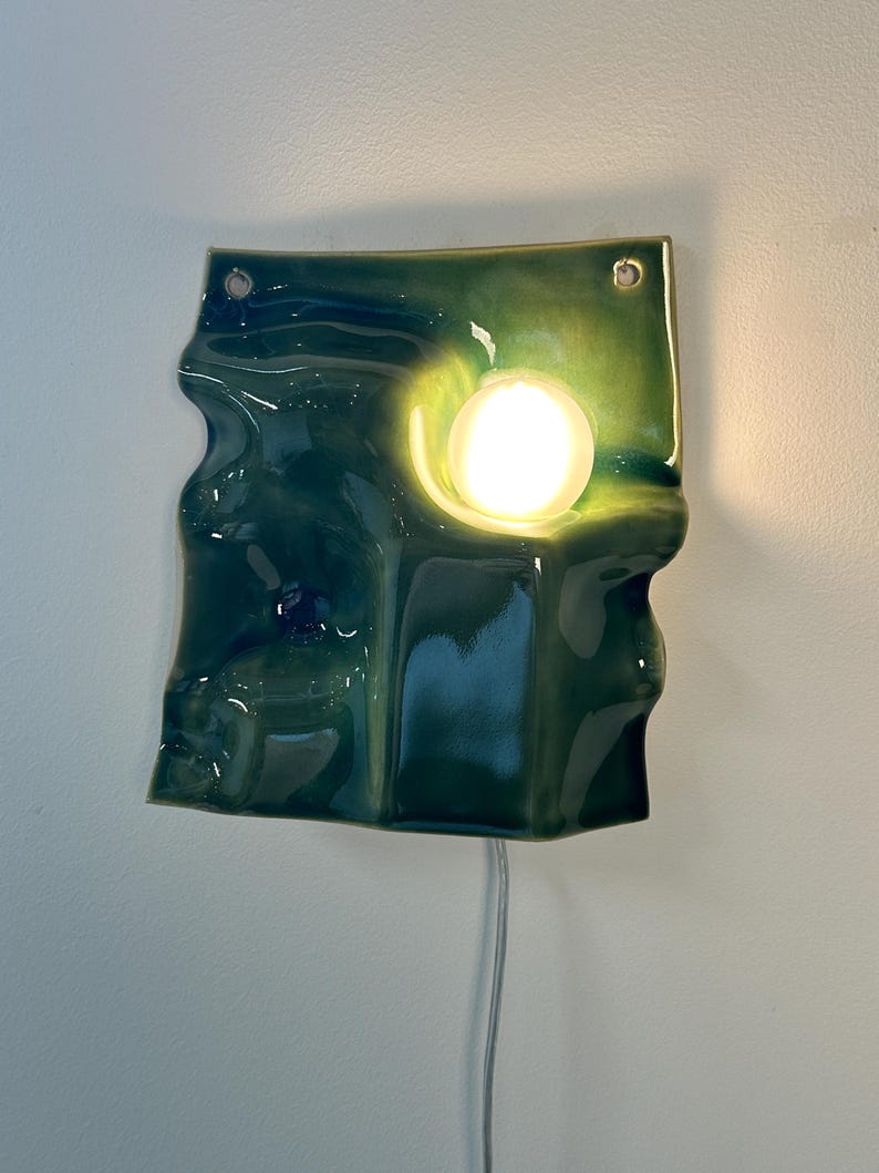 May include: A wall-mounted ceramic light fixture with a glossy, deep green, undulating surface. The square-shaped piece features a circular cutout holding a glowing, spherical white light bulb. A thin white power cord extends from the bottom edge.