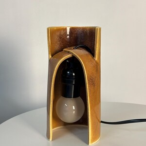 Puede incluir: Una l&aacute;mpara de mesa con una base de cer&aacute;mica cil&iacute;ndrica y curva con un esmalte brillante de color &aacute;mbar y marr&oacute;n moteado. La estructura tiene una abertura arqueada que alberga un casquillo negro y una bombilla blanca esmerilada con cable.