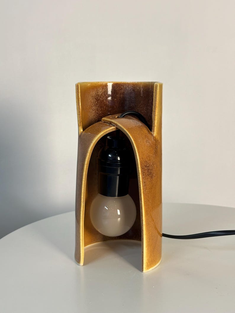Puede incluir: Una l&aacute;mpara de mesa con una base de cer&aacute;mica cil&iacute;ndrica y curva con un esmalte brillante de color &aacute;mbar y marr&oacute;n moteado. La estructura tiene una abertura arqueada que alberga un casquillo negro y una bombilla blanca esmerilada con cable.