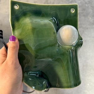 May include: A wall-mounted ceramic light fixture featuring a glossy, deep forest green glaze with a marbled finish. The rectangular piece has two circular mounting holes at the top and a single, round, frosted white glass bulb integrated into the front.