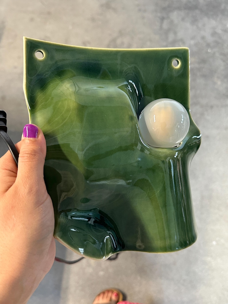 May include: A wall-mounted ceramic light fixture featuring a glossy, deep forest green glaze with a marbled finish. The rectangular piece has two circular mounting holes at the top and a single, round, frosted white glass bulb integrated into the front.